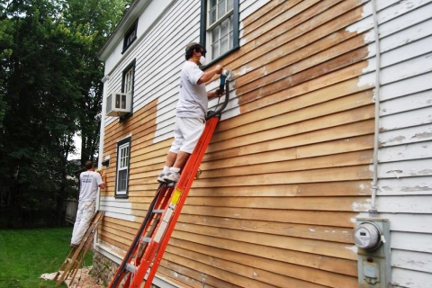 People painting the exterior wooden panels on a two story house.